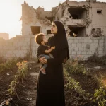 Young Palestinian woman hugging her child with wilted garden and a damaged building in background