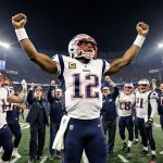 Drake Maye celebrates after a Patriots win with arms raised and teammates cheering near the Patriots logo in stadium lights