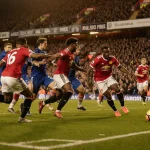 Manchester United player celebrating a goal with teammates from both teams surrounding the ball under light at Old Trafford