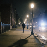 Streetlight casting long shadows on deserted sidewalk with a blurred man walking away in distance and police car passing.