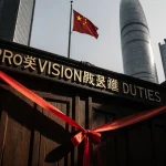 Ornate wooden gate displaying a red ribbon with a sign reading PROVISIONAL DUTIES above it near office towers and EU flag.