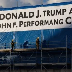 Banner displays memorial name with blue sky and workers building below.