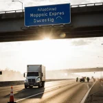 Truck drives southbound under overpass with reopening sign and emergency responders in background