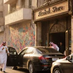 Cars queue outside an upscale liquor store with ornate facade and wooden sign while foreigners holding permits sip drinks.