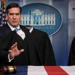 Secretary Marco Rubio gesturing toward a wooden table with a gavel and faded flag backdrop