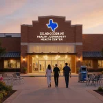People walking toward new community health center in Texas with golden light spilling inside and vibrant flowers around.