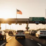 Cars speeding through San Ysidro border lanes with Mexican and American flags and a warm sunrise glow