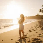 Savannah Louie standing on a beach with arms crossed and a smile as sunset lights her profile over golden sand palm trees
