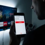 User peeking over smart TV with smartphone screen showing red Tracking notification and ominous LED glow