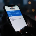 Hands gripping smartphone displaying urgent warning alert with dark morning cityscape and blurred car in background