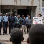 South African authorities standing at entrance of refugee center with Kenyan detainees escorted by officers and protest signs