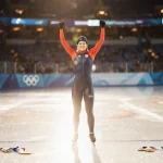 Aaron Jackson triumphantly raising arms at speed skating finish line with reflected ice lights and gold medals near her