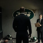 Steve Wilks stands alone in dim locker room with faded Jets logo and scattered footballs and helmets.