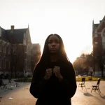 Student standing alone before Brown University Gothic buildings with clenched fists and shadowed face under sunset light.