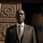 Sudan Prime Minister stands before an ornate wooden door ajar with warm light and a blurred African map in background