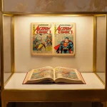 Golden-lit display case illuminates open vintage Superman No. 1 comic with stacks of old comics and warm beige background.