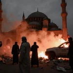 Alawite protesters stand defiantly with smoke and flames from burning vehicles and a mosque silhouette in the background