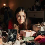 Teenage girl scrolling on cracked phone with a meh look and unwanted gifts scattered around her and a gaming console beside a