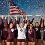 Texas A&M volleyball team celebrates with arms raised and confetti falling as Kansas City skyline Missouri River behind flag.