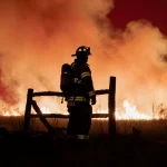 Firefighter silhouette standing and gazing at distant wildfire with broken fence post and crimson Texas sky