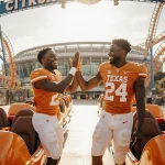 Two Texas Longhorns players laughing and high‑fiving with colorful roller coaster cars and Orlando amusement park backdrop