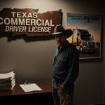 Truck driver looking concerned with crooked wooden sign and abandoned grant documents near a faded semi‑truck poster.