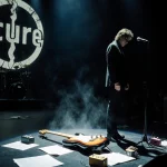 Robert Smith standing at microphone with abandoned guitar and mist, The Cure logo backdrop