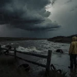 Lone figure standing at riverbank watching turbulent water with stormy clouds and a wooden fence in background.