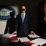 Todd Blanche standing before a wooden desk with redacted files and a faint Trump logo behind him near a courtroom doorway