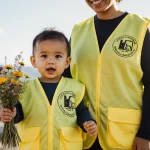 Toddler holding wildflowers with parent in matching yellow vests against a blurred Denver cityscape for a community garden.