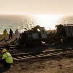 Worker kneels beside mangled railcar with twisted metal and hazmat searchers on desolate rail line with Pacific Ocean horizon