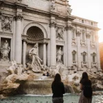 Tourists gather near Trevi Fountain with warm dawn light and coins scattered on stone.
