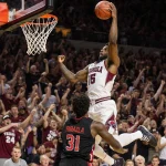 Trevon Brazile leaps over Texas Tech bench dunking a ball with Arkansas jersey logo amid blurred crowd of fans.