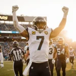 Trevor Lawrence celebrates two touchdowns with arms raised while teammates surround him beneath Jags logo and golden sunset.