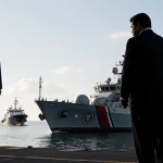 President Trump standing beside black Coast Guard cutter with stern pointing toward horizon and silhouette of Maduro dock.