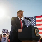 President Donald Trump speaking at podium with American flag and cheering crowd under blue sky