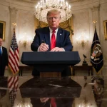 President Trump holds a pen near the White House podium with chandeliers and marble floors below and flag blurred behind