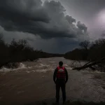 Person in life jacket standing at swollen Upper Guadalupe River with lightning-lit sky and uprooted trees