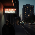 Two people huddling together with neon Emergency Shelter sign and dimly lit urban alley in background.