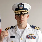 Coast Guard officer stands proudly with hand on U.S. emblem near American flag and soft neutral background.