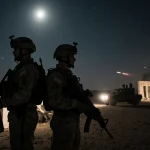 US soldiers standing back‑to‑back near a makeshift command center with moonlit desert and distant artillery fire