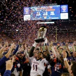 Virginia players hoisting the championship trophy with ecstatic fans waving confetti and a glowing scoreboard in background