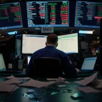 Trader sits hunched over computer with scattered papers and shattered glass on a dimly lit Wall Street trading floor