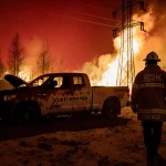 Firefighter walking away from wildfire with charred trees and flames licking a power line and abandoned rusted truck