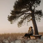 Person sits on wooden bench with cozy light and a majestic cedar tree in a snowy winter landscape.