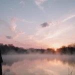 Lone figure standing at lake edge gazing east with sunrise glow and mist over winter morning lake