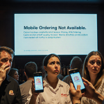 Frustrated bar staff at Alamo Drafthouse stare at glowing error screen with customers holding phones in disappointment.
