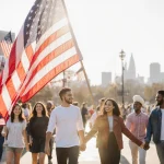 American flag waving in front of community center with diverse young volunteers holding hands and smiling in sunlit patriotic