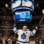 Auston Matthews raises his stick in celebration with confetti and the Toronto Maple Leafs logo in the background