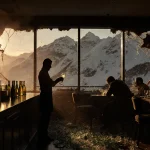 Bartender standing amid shattered champagne bottles with snowy mountains and fire remnants in background
